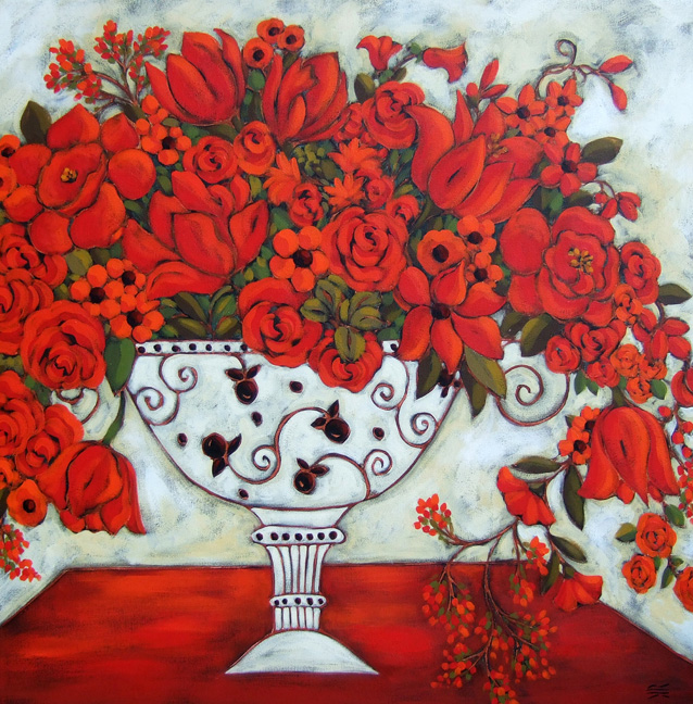 A White Jardiniere with black floral patterning is filled with an abundance or red flowers including tulips and roses. the background is white and the vase sits on a brilliantly coloured red table.